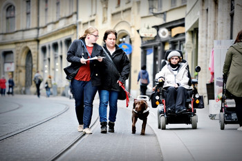 Frau im Rollstuhl, Frau mit Blindenhund und Frau mit Schreibutensilien sind in der Stadt zusammen unterwegs
