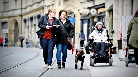 Frau im Rollstuhl, Frau mit Blindenhund und Frau mit Schreibutensilien sind in der Stadt zusammen unterwegs