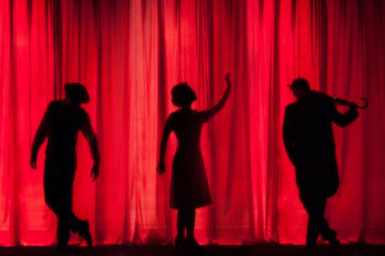 Schwarze Silouetten von Darsteller:innen auf einer Bühne mit rotem Vorhang dahinter