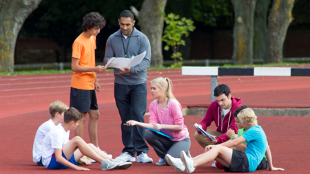Gruppe junger Menschen auf einem Sportplatz mit ihrem Trainer: 5 sitzen auf dem Boden, ein Kind spricht mit dem Trainer